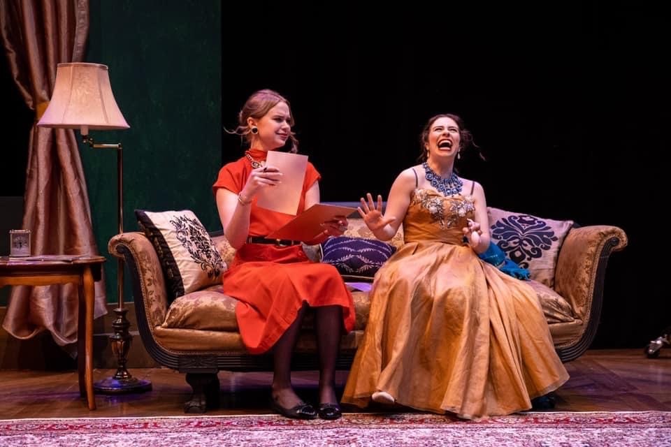 two actors on a couch in costume, one with a paper and one laughing