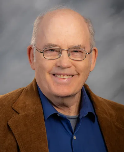 professional headshot with Tom Paradise smiling, wearing glasses, dressed in a brown leather jacket and navy blue collared shirt