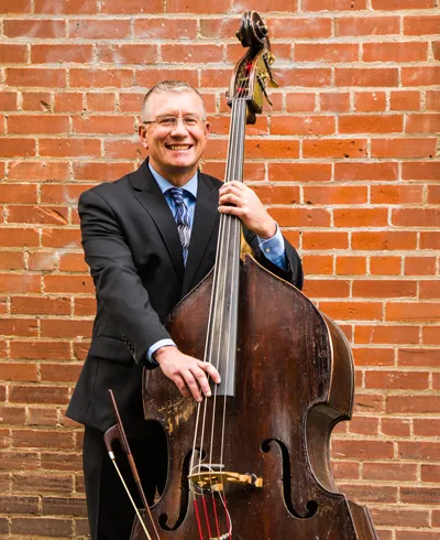 Jason Crowe in a 3-piece suit holding an upright bass in front of a red brick wall