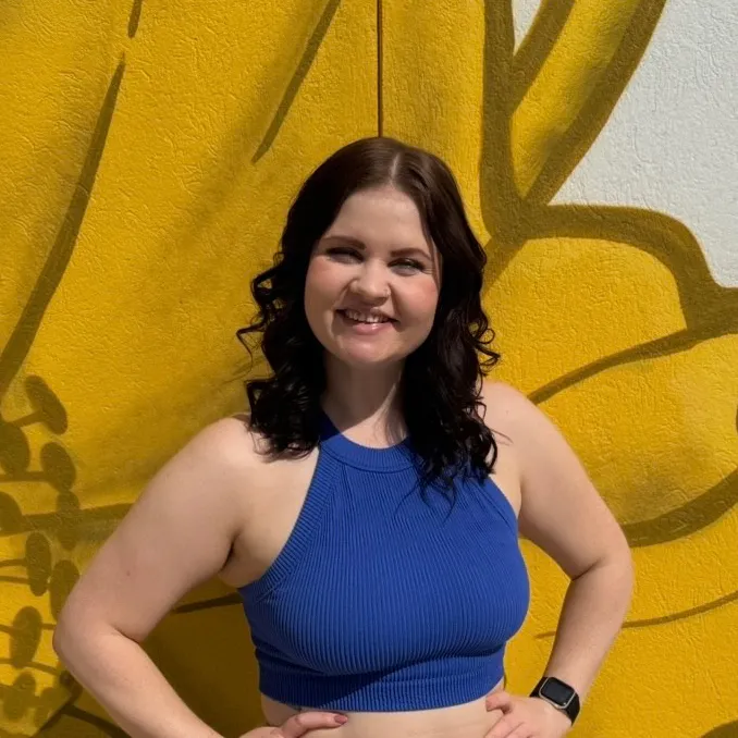 woman with dark curly hair standing in front of yellow flower mural, arms bent out at sides, smiling