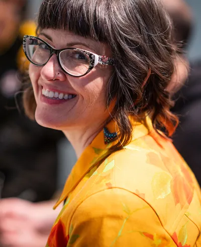 Joy Armstrong with glasses and yellow coat looking back over left shoulder smiling at camera